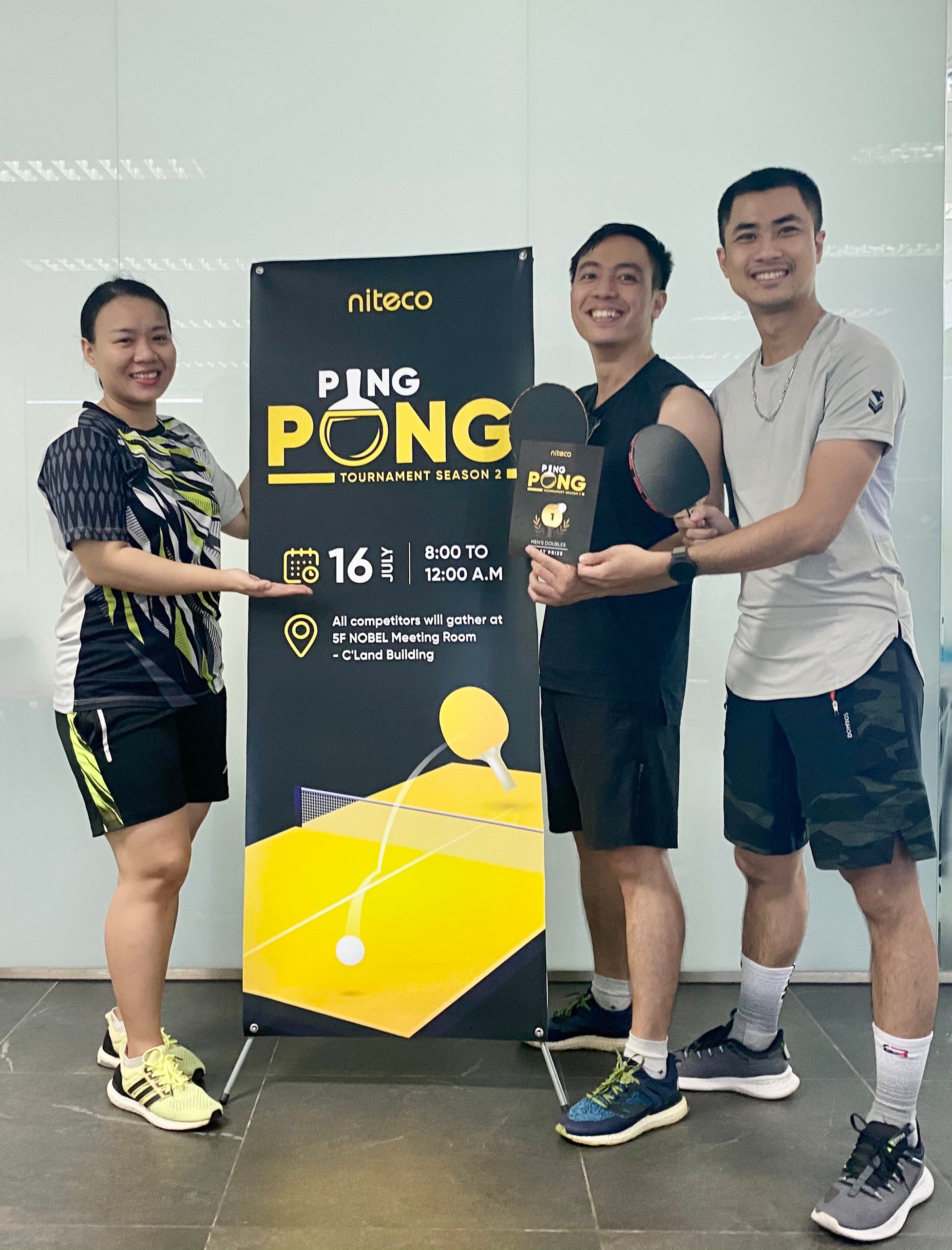 Three players smiling with ping pong paddles and a tournament poster.