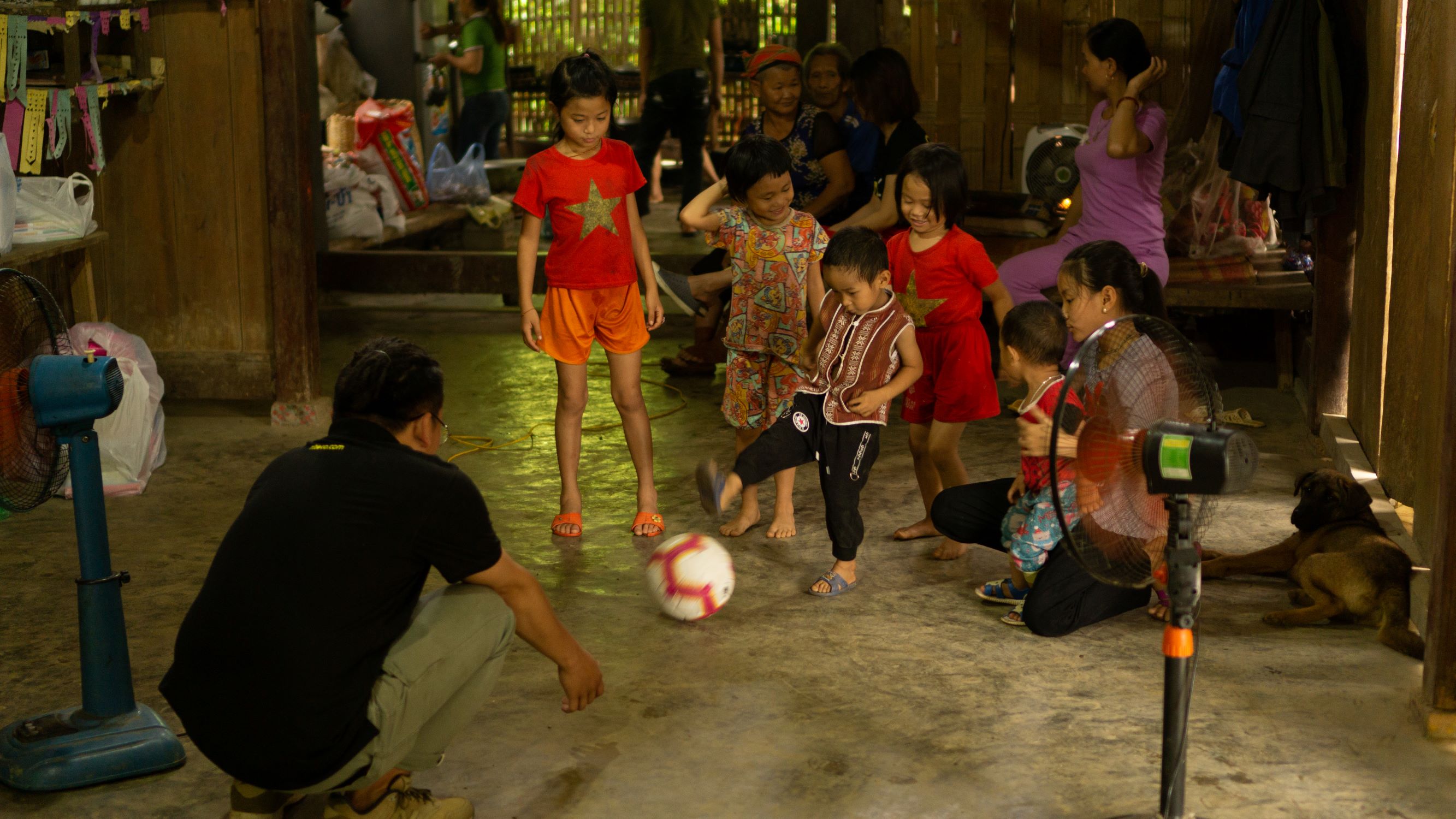 Children playing indoor football with family and a pet dog around fans