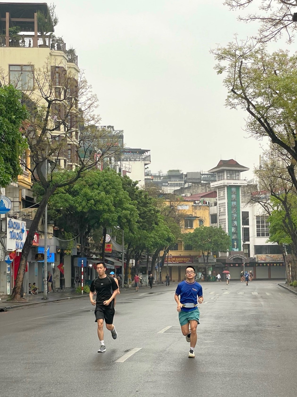 Two young men jogging on a quiet Hanoi street lined with trees and buildings