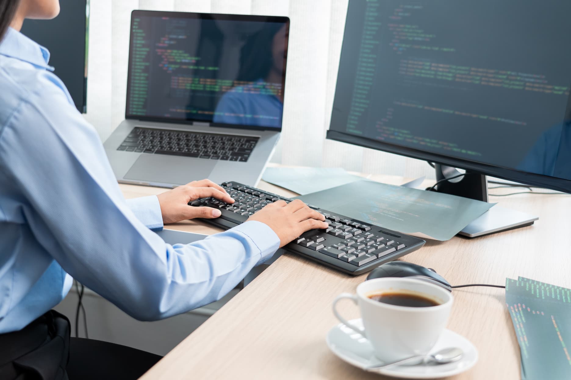 Person coding on dual monitors with coffee cup nearby on desk