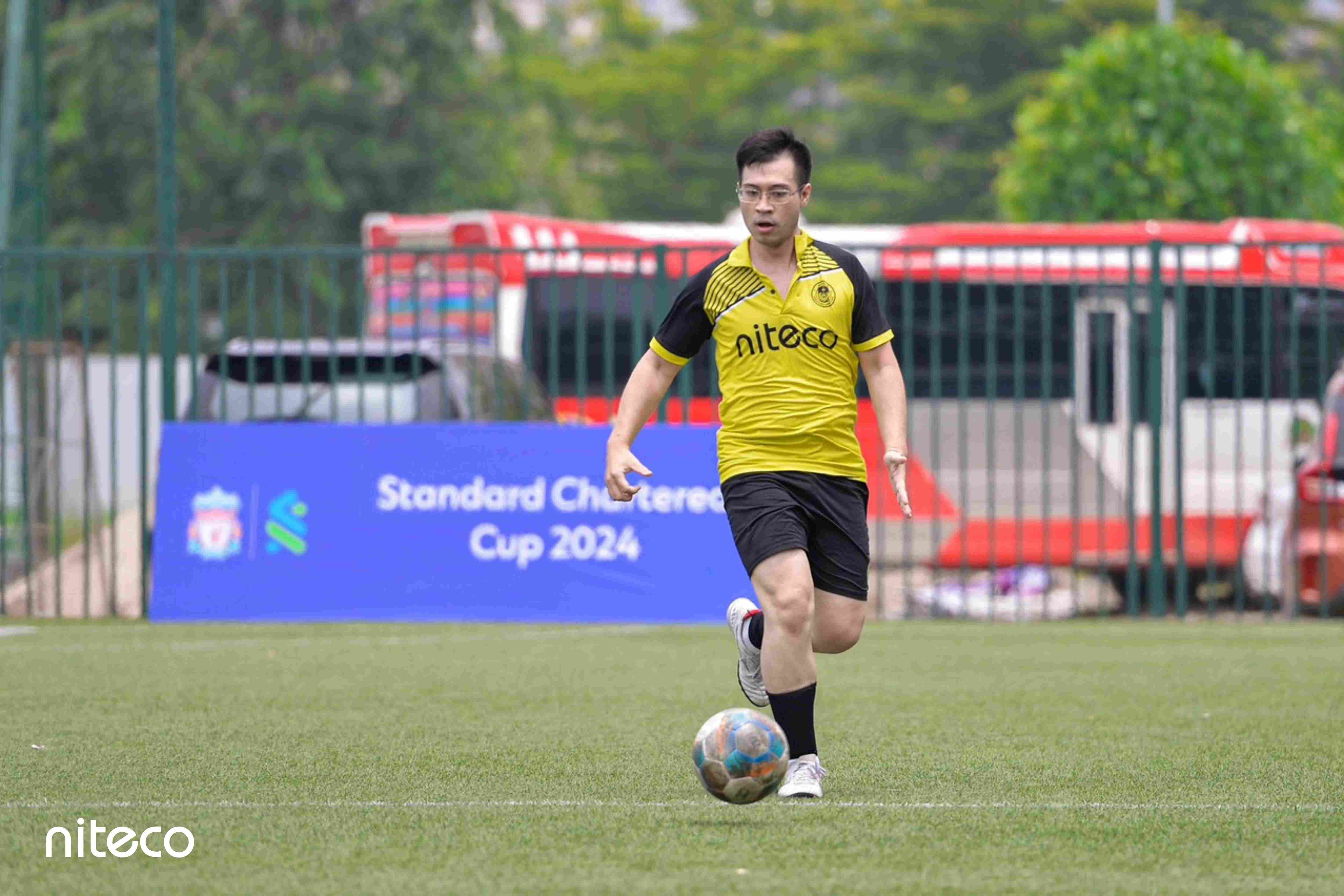 Football player in yellow Niteco jersey dribbling ball at Standard Chartered Cup 2024