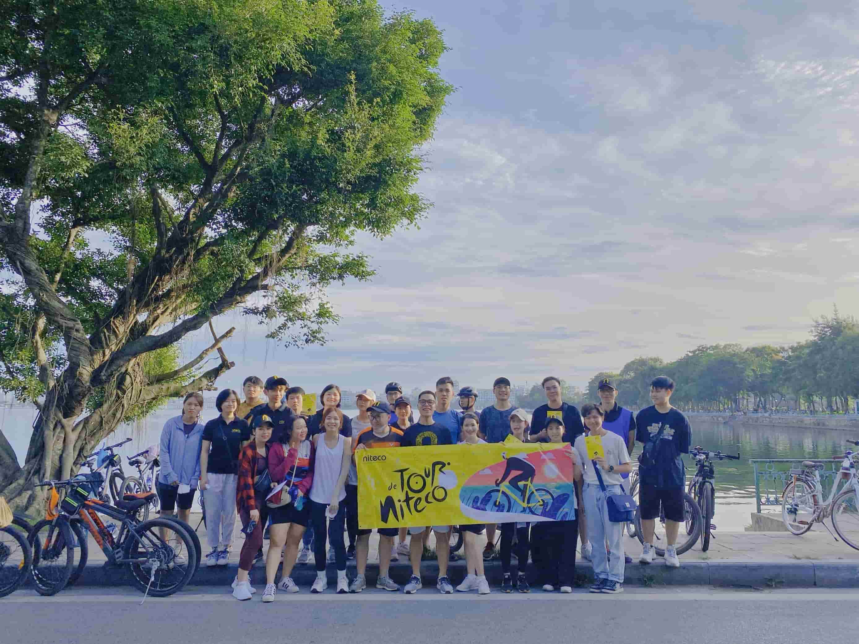 Group of cyclists holding Tour Niteco banner by lakeside with bikes and trees