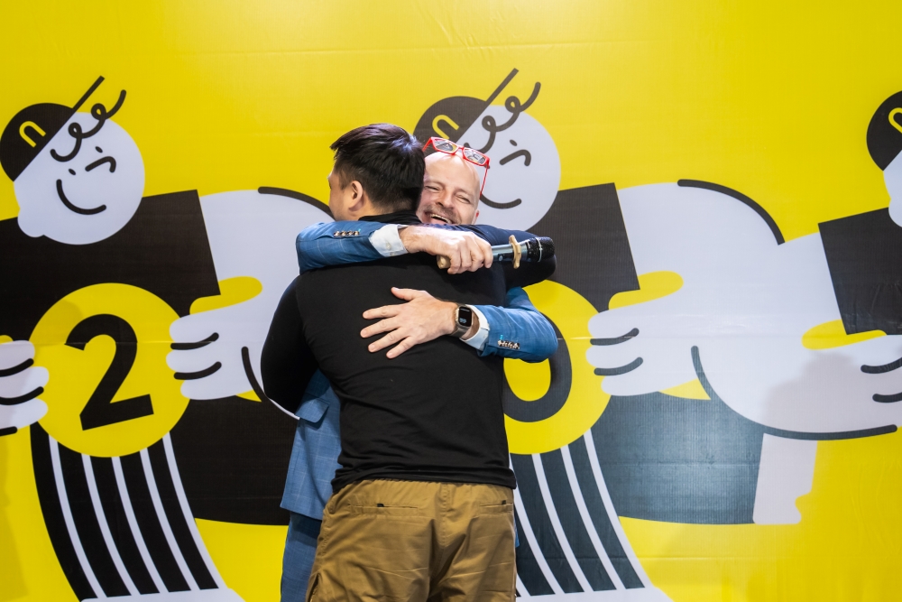 Two men hugging warmly at the 2020 event with bright yellow background