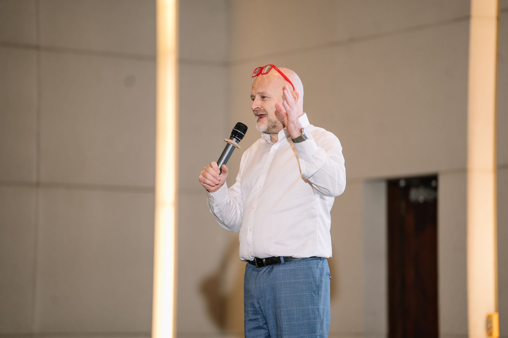 Bald man with red glasses speaking on stage holding microphone during presentation