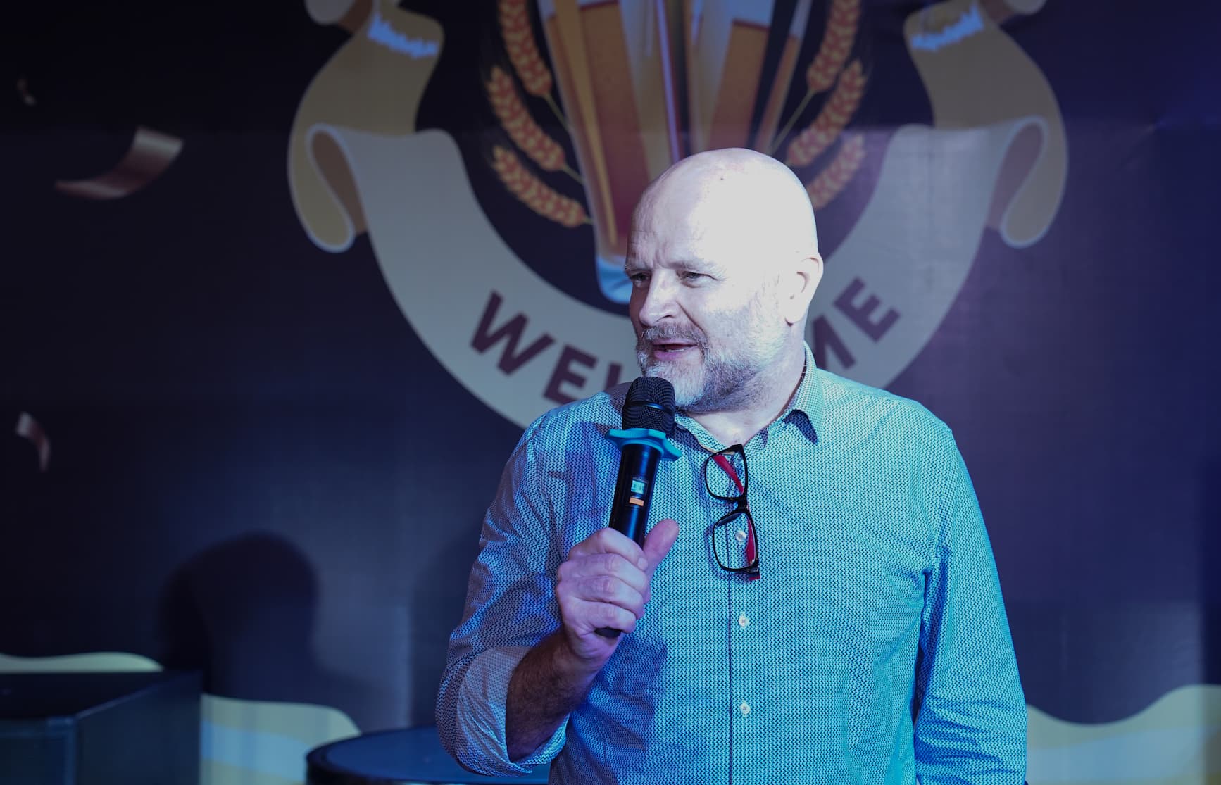 Man holding microphone speaking at a welcome event with wheat emblem background