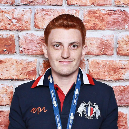 Red-haired man wearing navy polo with crest and event lanyard against brick wall