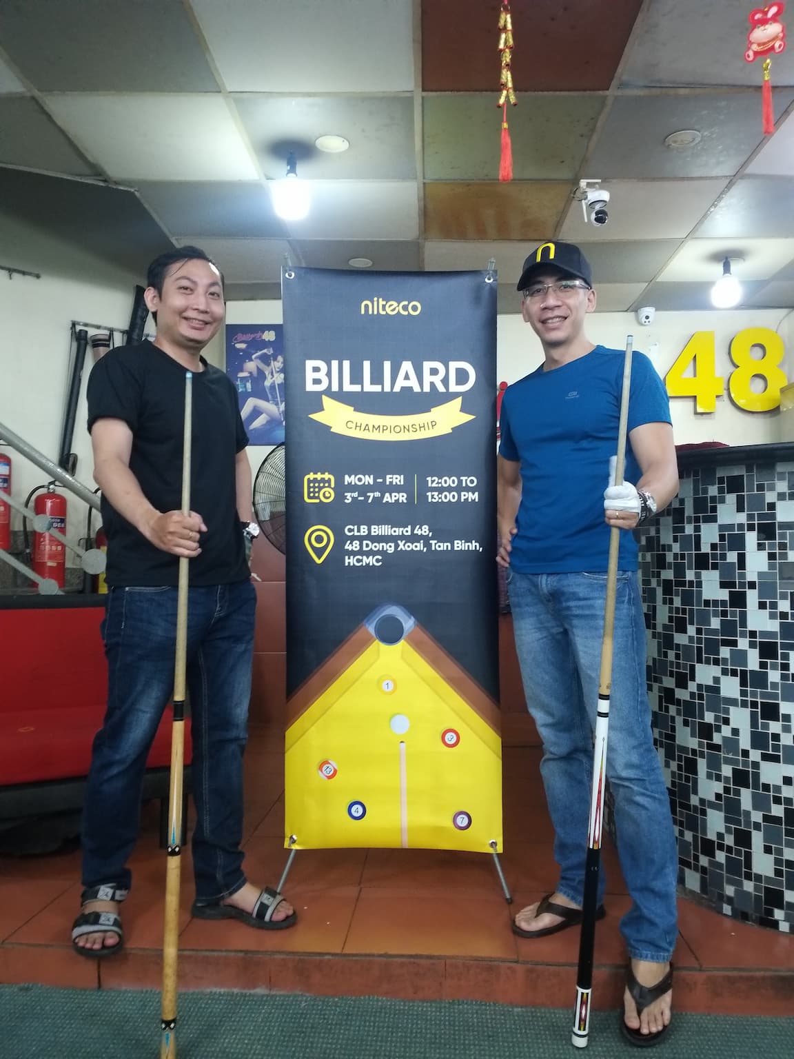 Two men holding billiard cues stand beside a banner for Niteco billiard championship event.