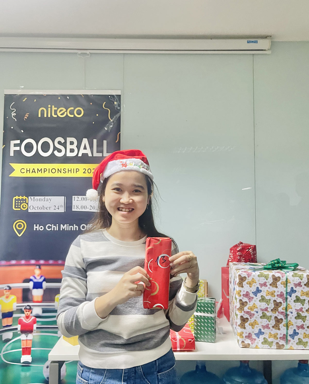 Woman wearing Santa hat holding gift near Foosball Championship 2022 poster